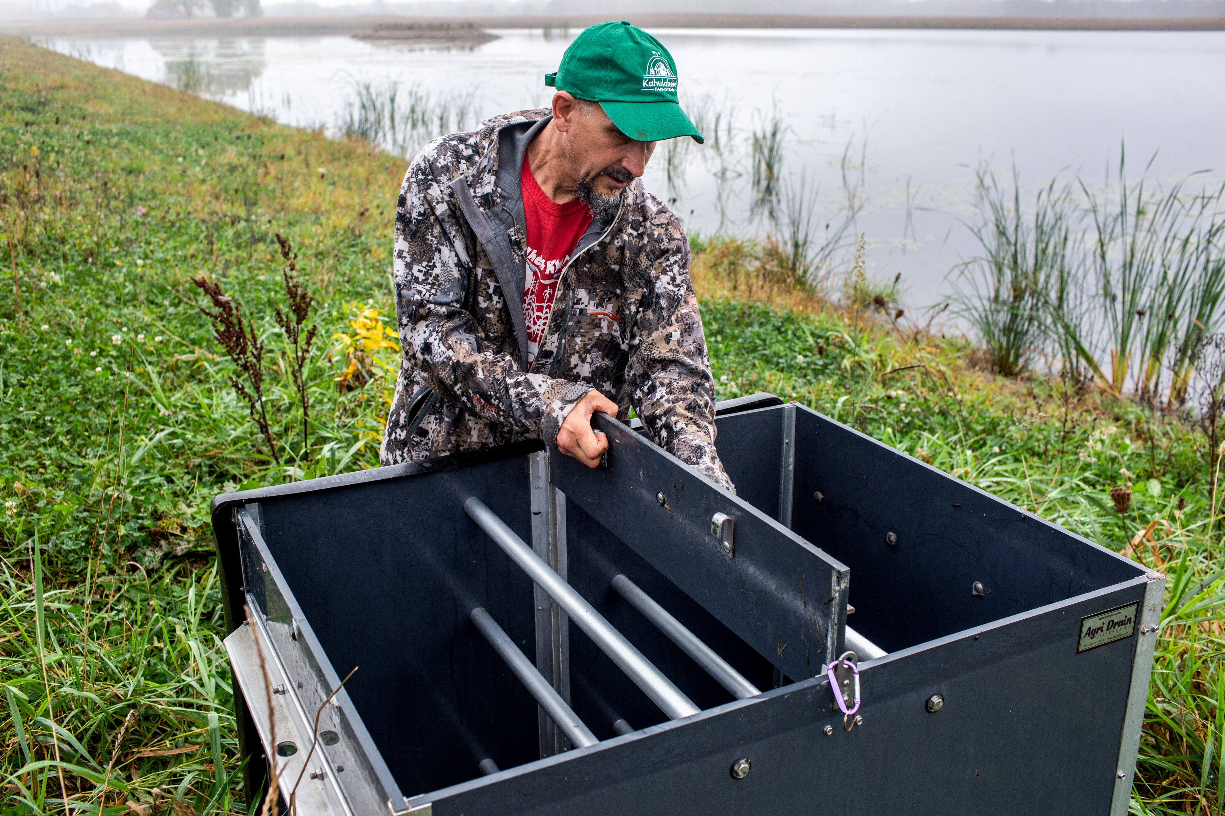 Tony Kuchma, a man wearing a green baseball cap and a red t-shirt under a camouflage patterned jacket, checks water levels in a big, dark-blue metal box that is foregrounded in the photo. In the background are green reeds and a stretch of flat, blue water—the restored wetlands of the Oneida Nation in Wisconsin.