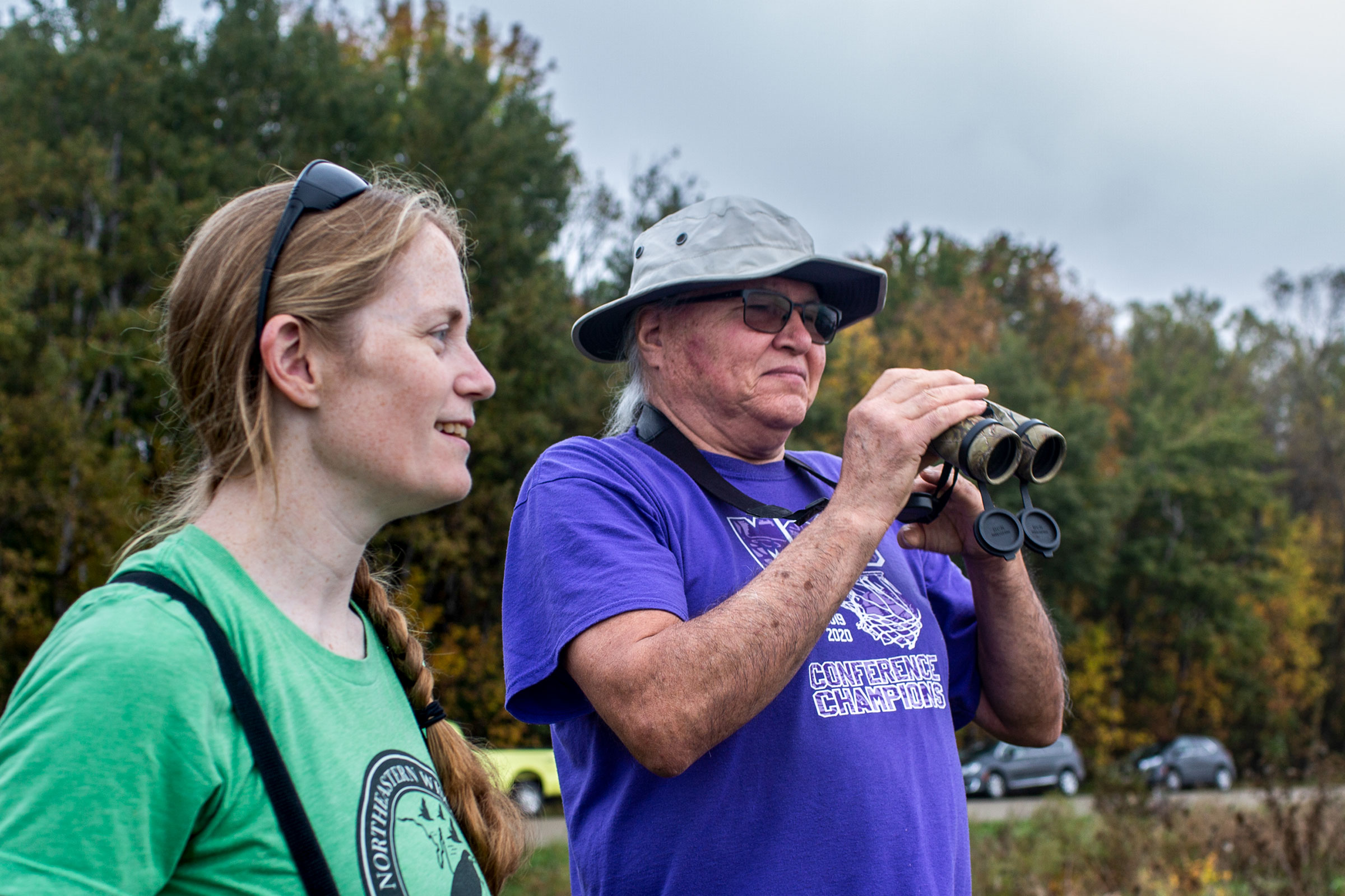 Erin Giese is wearing sunglasses and a green shirt, and standing to the left of Randy Cornelius, who wears a purple shirt and a white bucket hat and holding a pair of black binoculars in his hands. Both are looking to the right of the photo frame as they monitor birds in the Oneida Nation's restored wetlands.