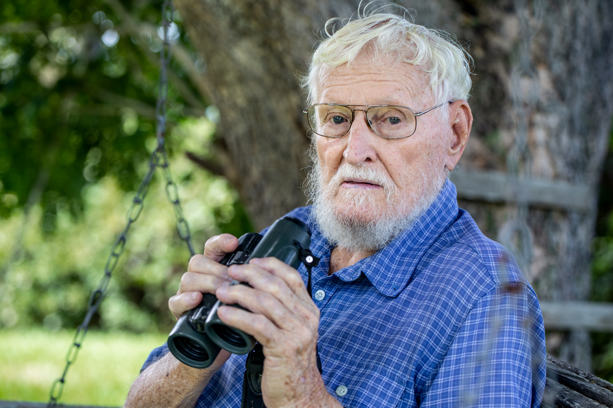 David Wingate poses for a headshot. He has white hair, a white beard, a bright blue button-down shirt, and grasps a pair of binoculars.