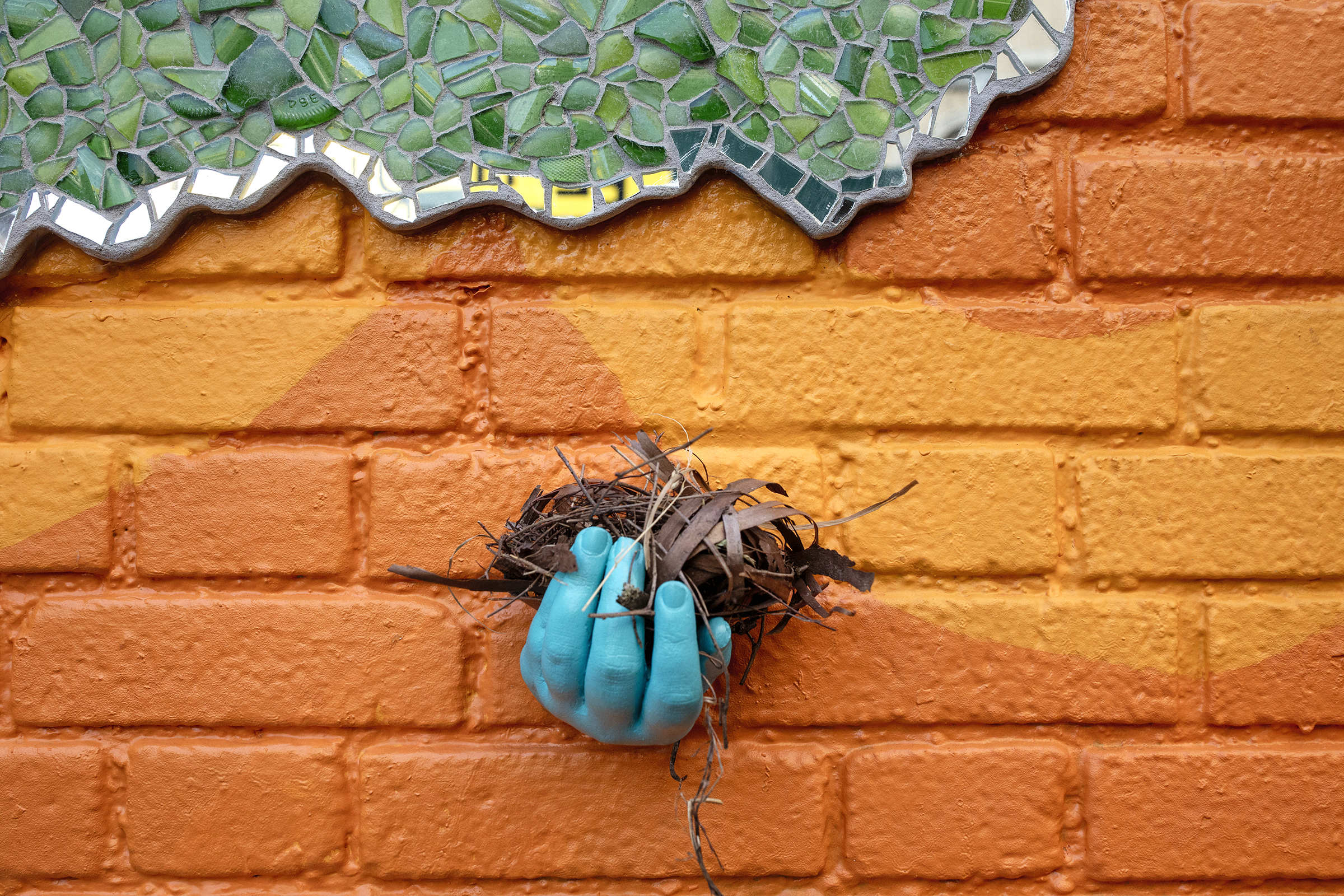 A concrete hand, painted robin's egg blue, juts out from a bright orange brick wall. Inside the hand is a small pile of brown twigs and bark resembling a birds nest. In the upper left quadrant, green and mirrored mosaic are visible.