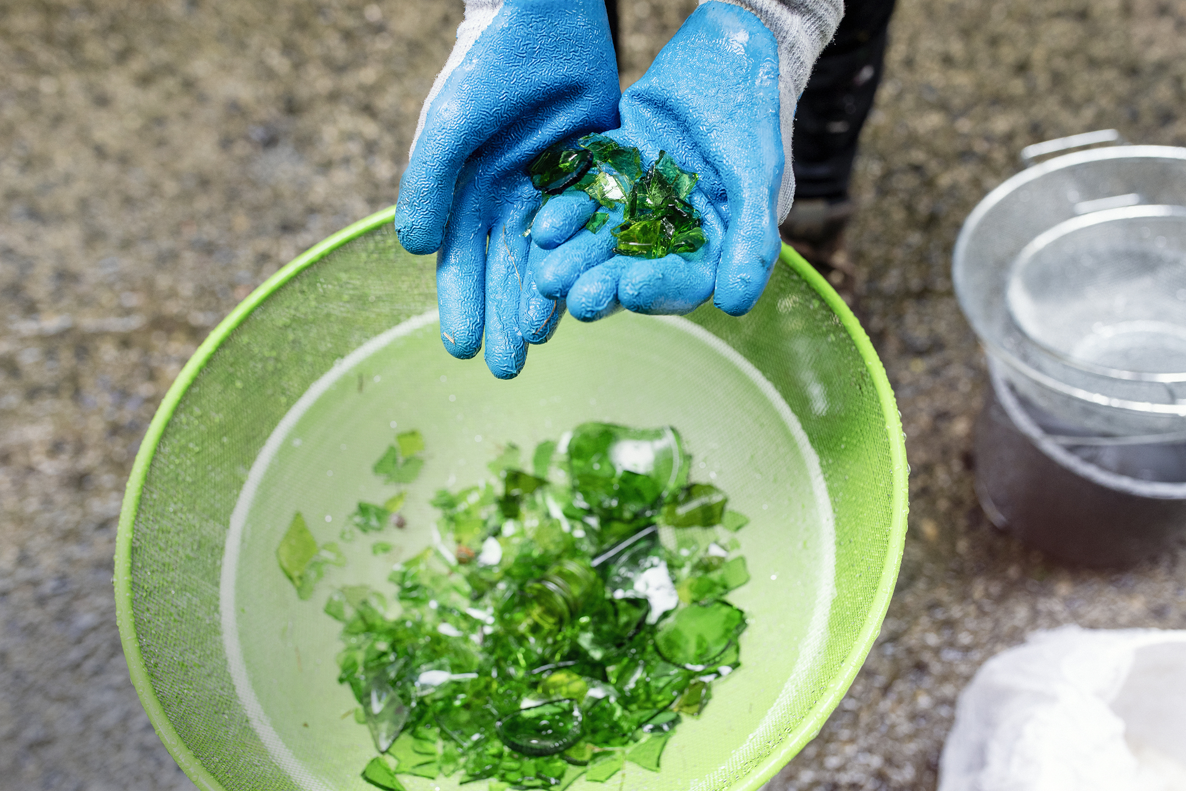 Hands wearing bright blue gardening gloves cup shards of emerald green glass over a lighter green pan.