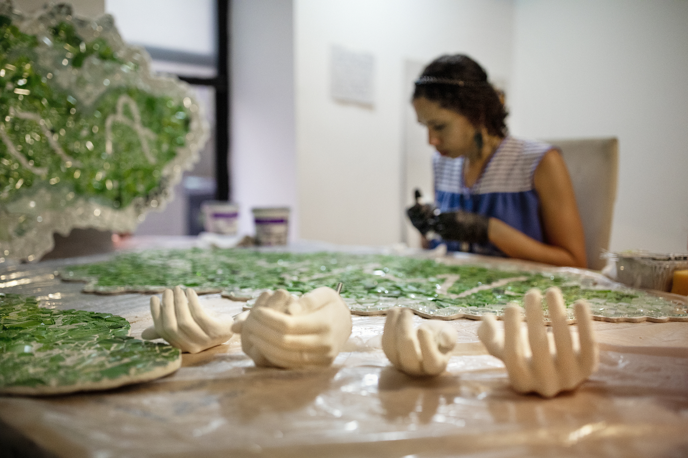 A woman sits in concentration at a long table in a white-walled room. In the foreground are sections of green and clear glass mosaic clouds and four hands cast from concrete.