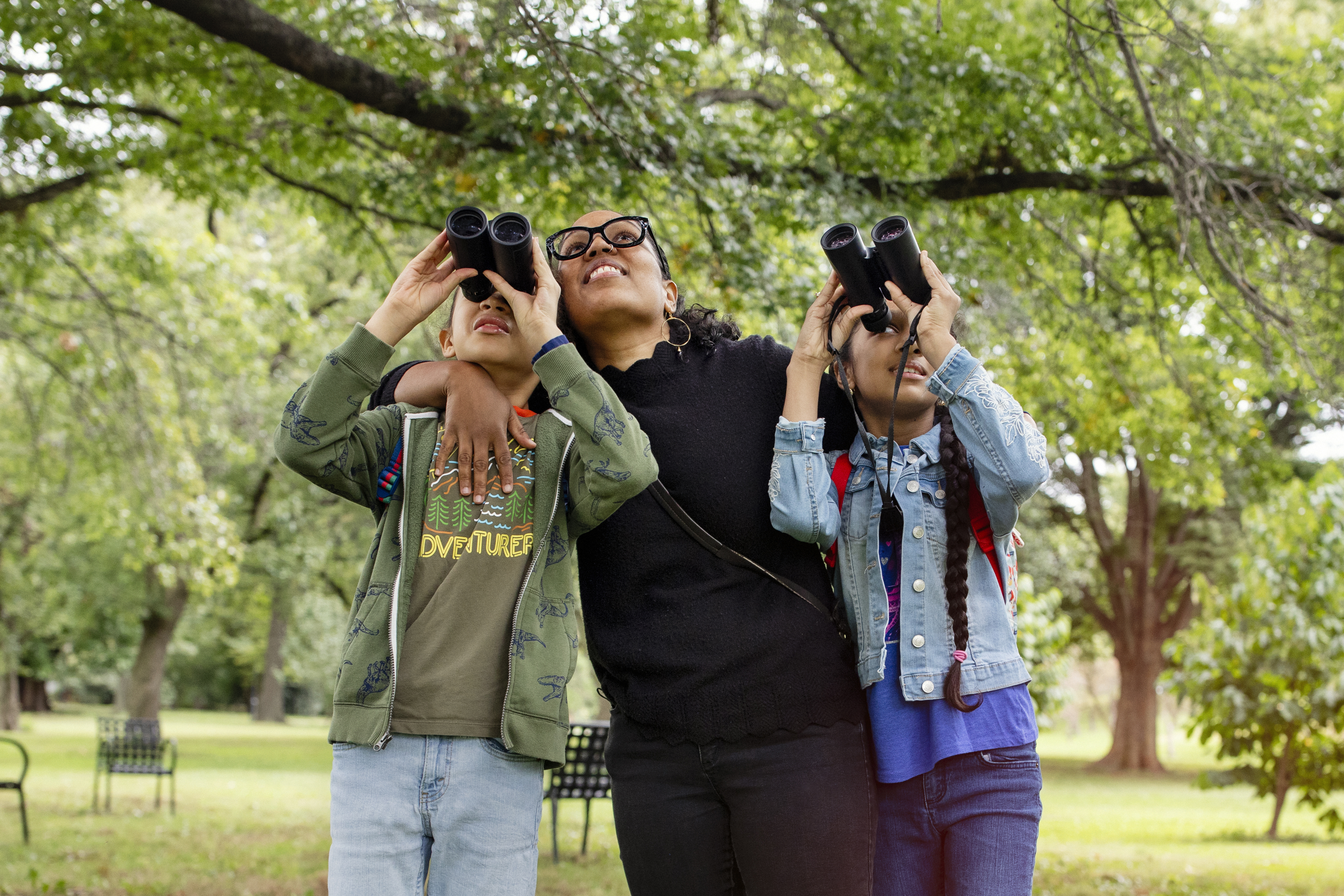 A mother stands between her children, her arms wrapped around them as they look through binoculars. They are in a park, with trees and metal benches behind them.