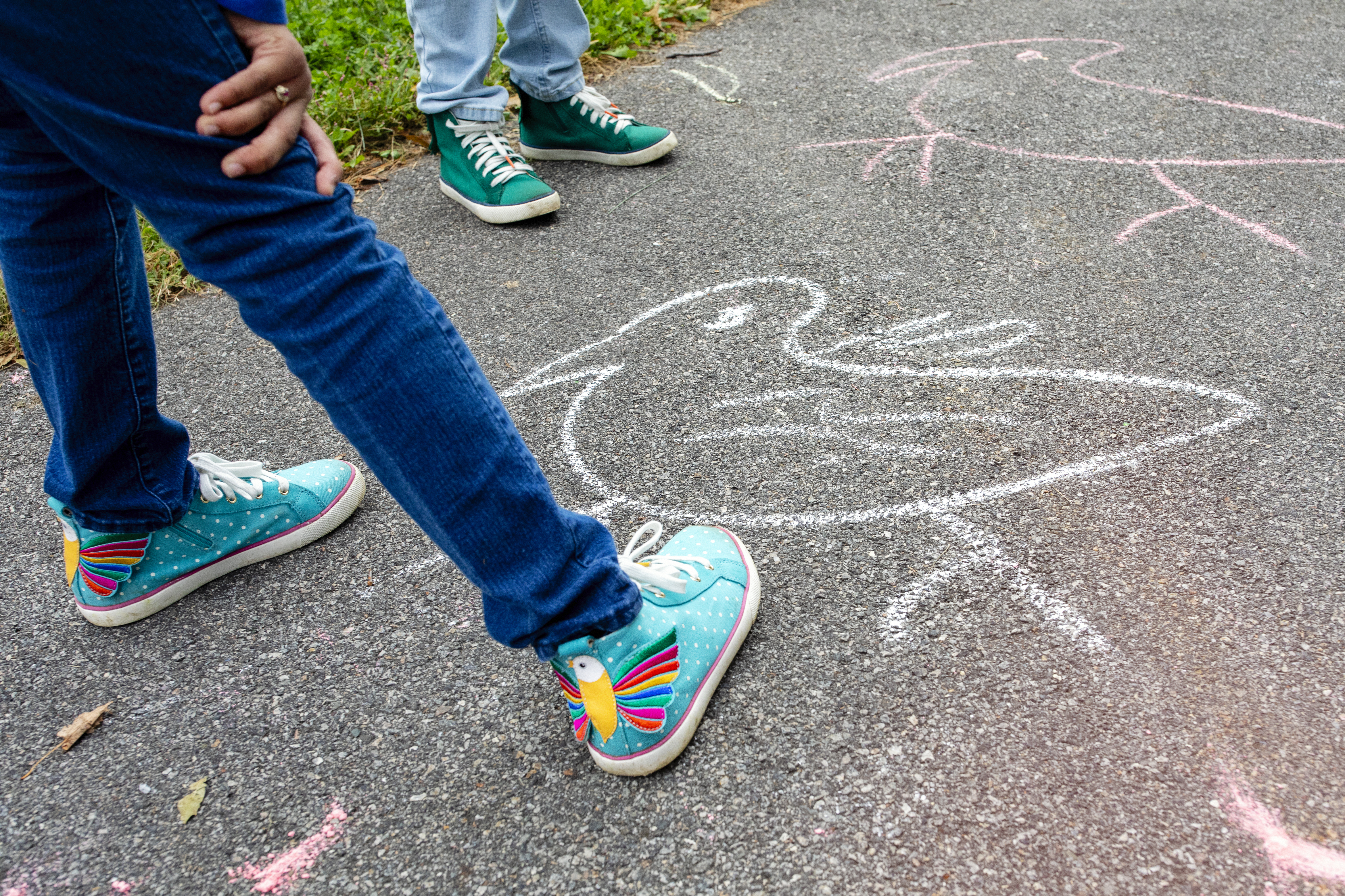 Two children, only their legs showing, stand on a bath looking at pink and white chalk drawings of birds. The shoes of the closest child boast colorful birds on the back, with yellow bodies and rainbow-patterned wings.