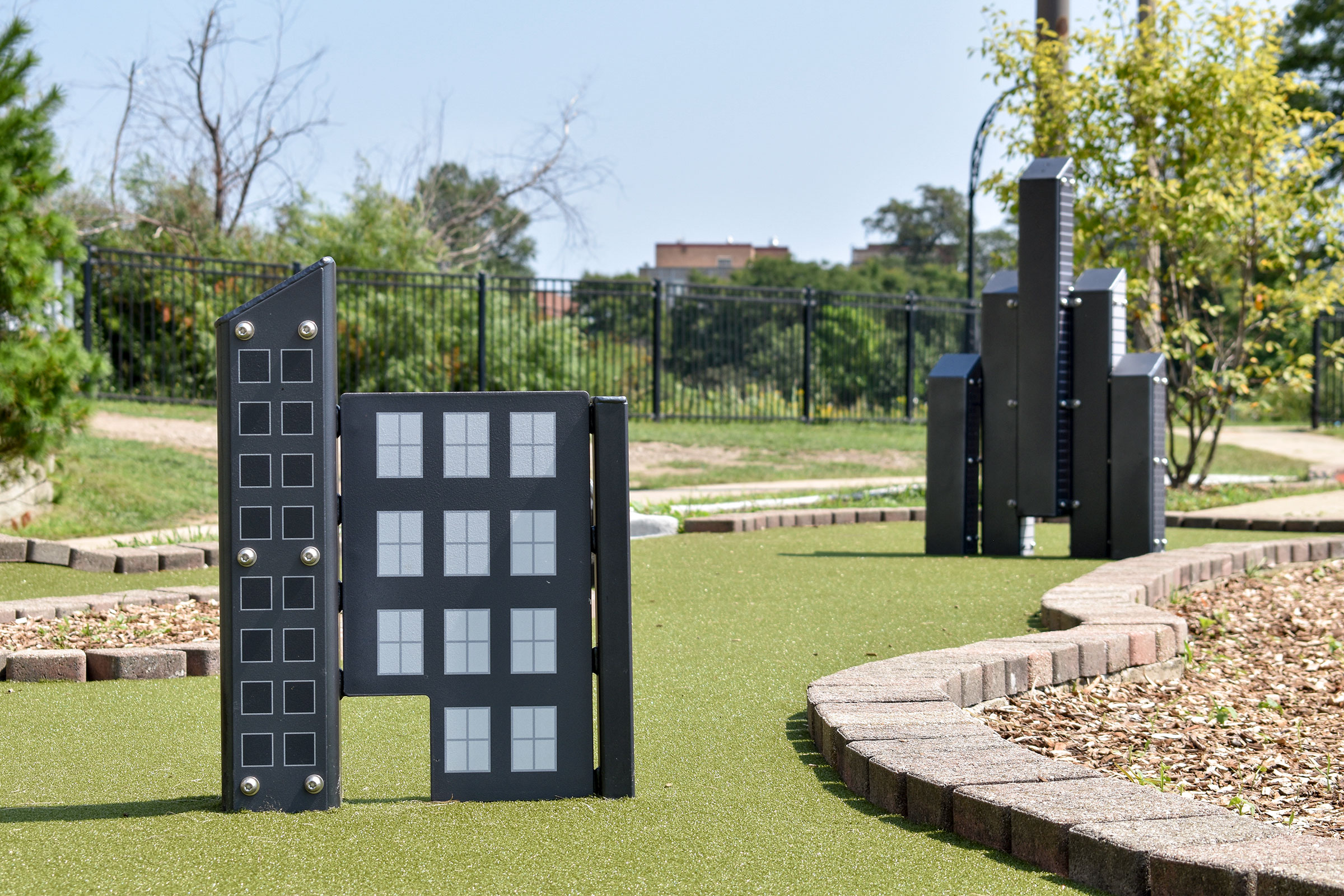Miniature buildings reminiscent of Chicago's skyscrapers stand out from green turf. Trees and buildings are in the background.
