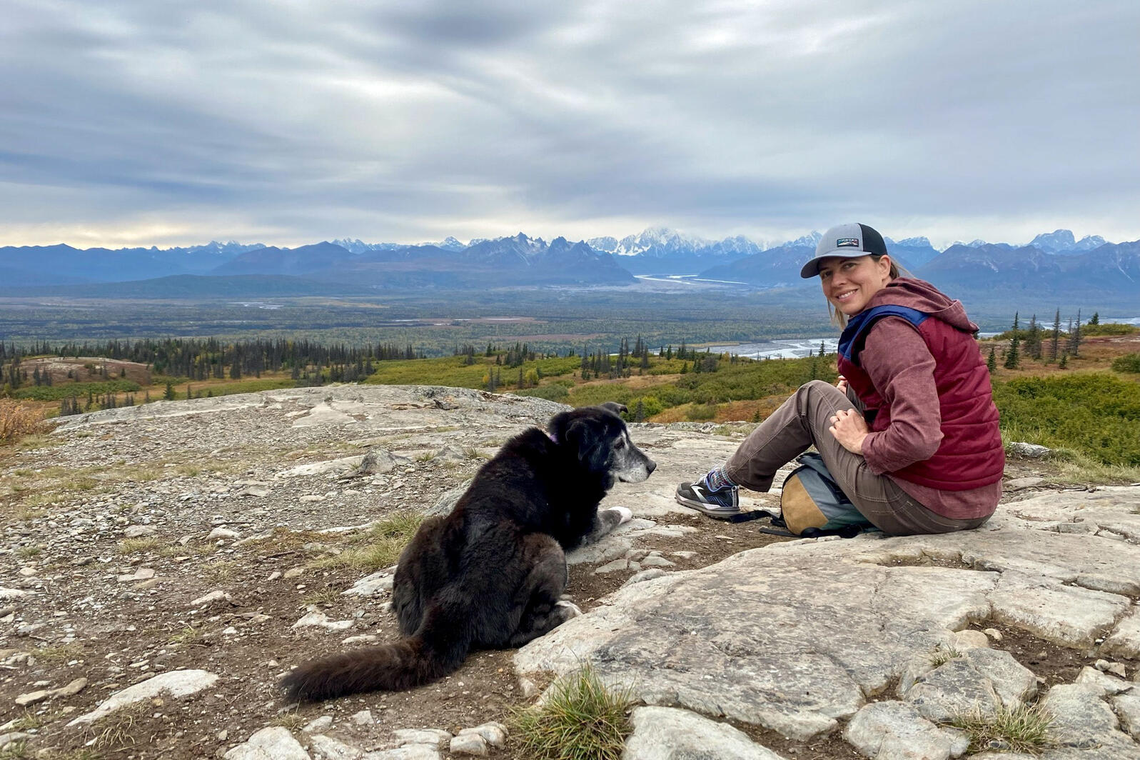 Melanie Smith and a black dog sit on a rock looking out over a lush green forest and distant, snow-capped mountains. 