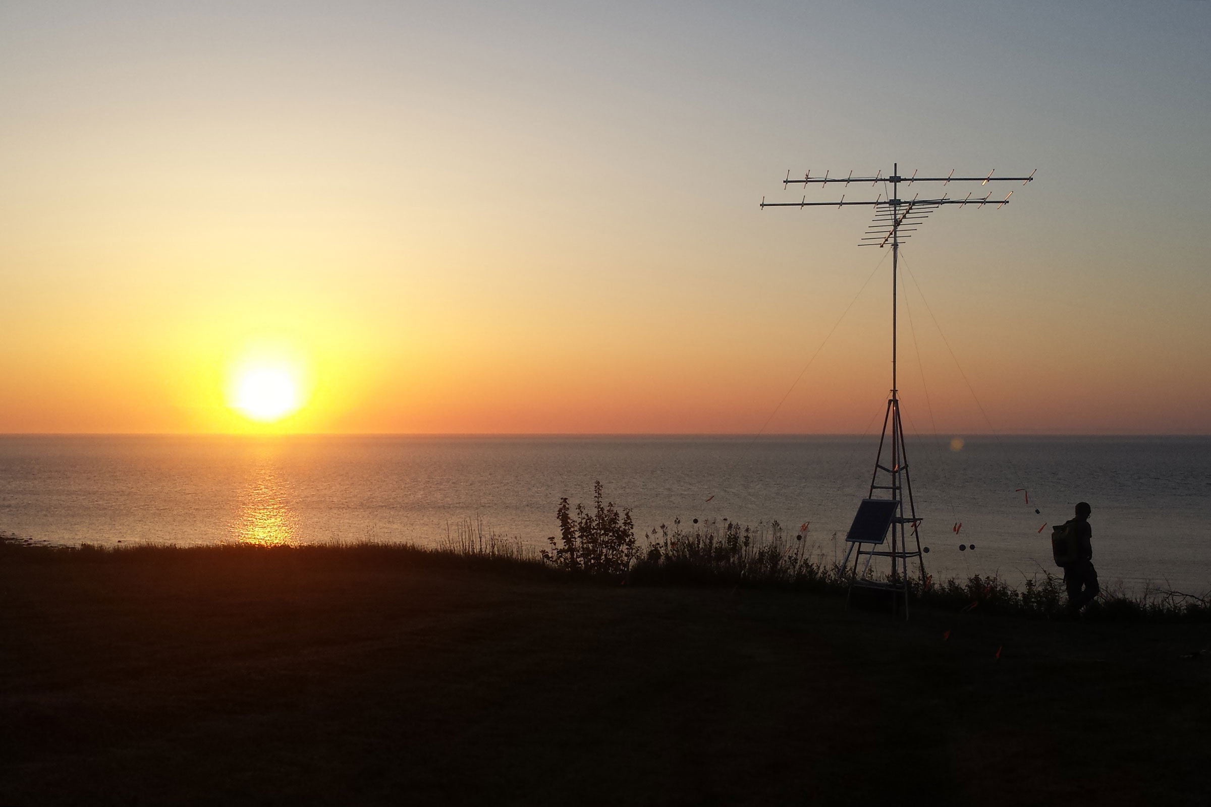 The sun sets over water, and a tall antenna is in the foreground.