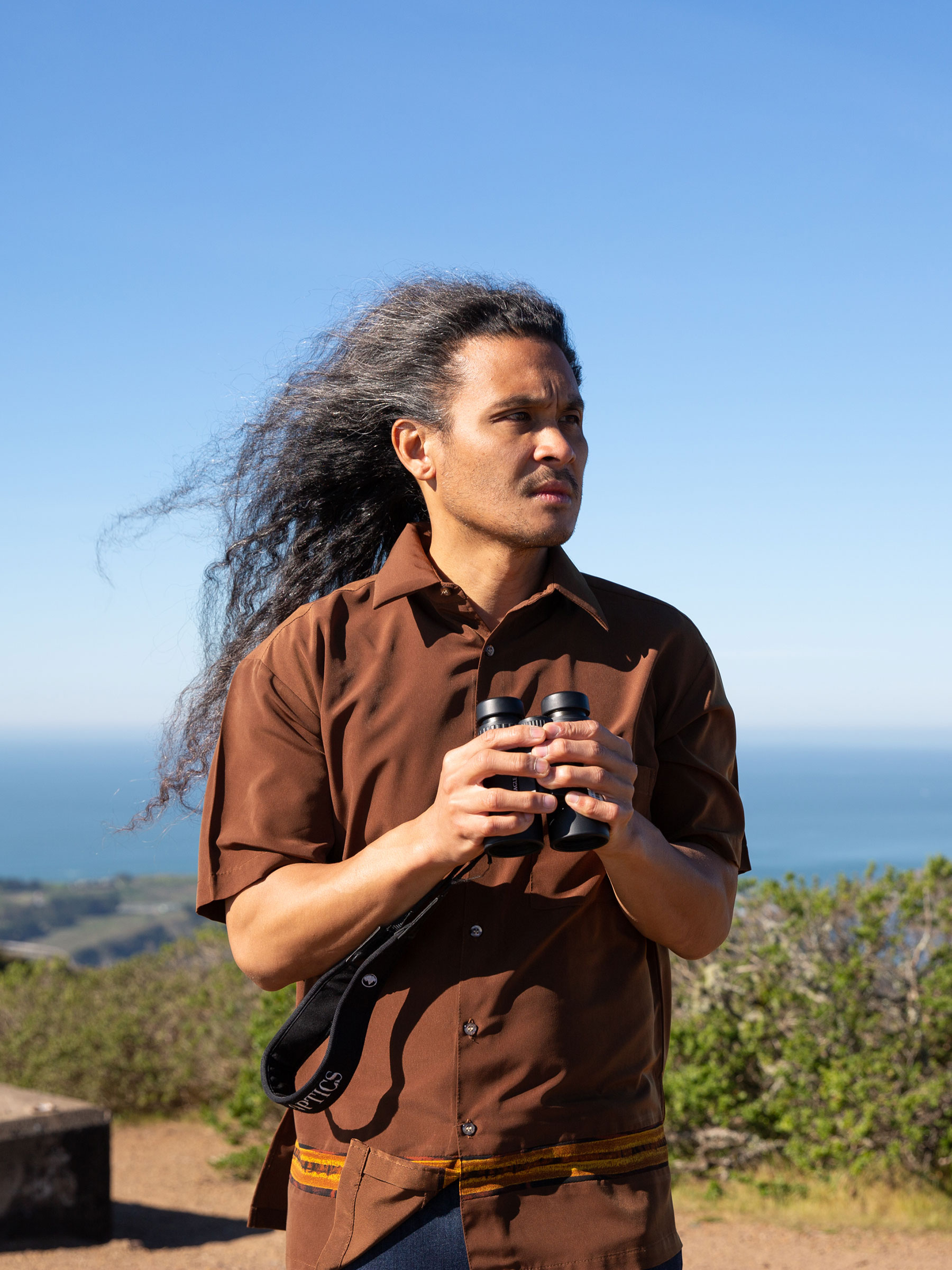 A man with long flowing hair stands on top of a hill, with water in the background, holding binoculars.