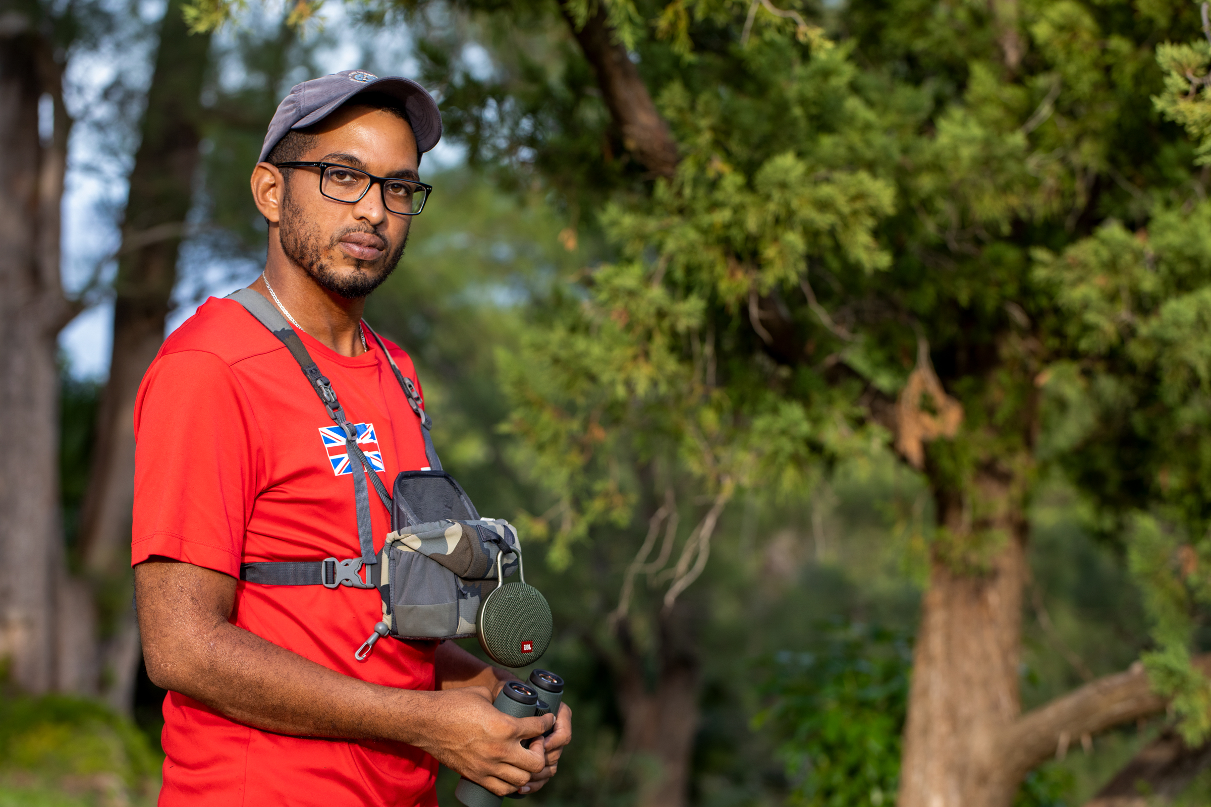 Miguel Mejías is a young man who could be the next Cahow caretaker. He stands for a portrait in front of greenery wearing a red t-shirt, baseball cap, and black-frame glasses. He holds a pair of binoculars.