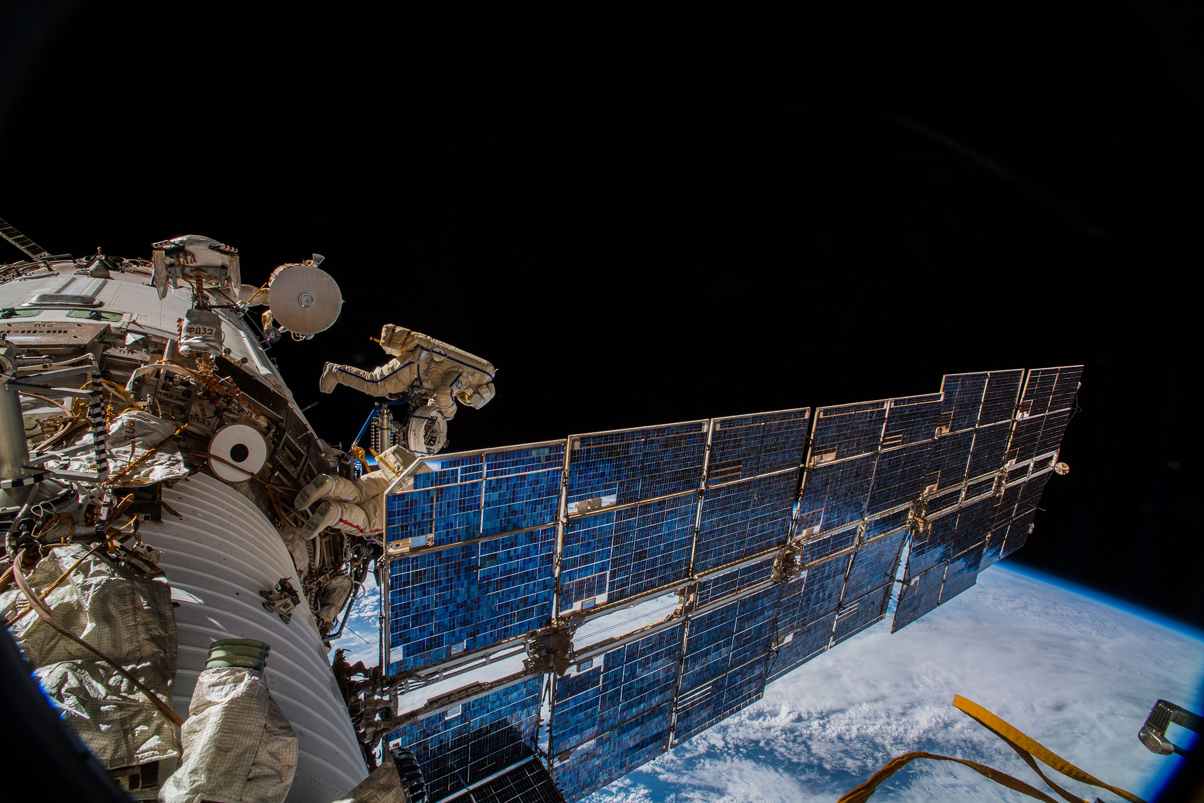 Oleg Artemyev and Sergey Prokopyek install an antenna for the ICARUS animal tracking software on the International Space Station in 2018. A large spacecraft with massive solar panels hovers with the Earth in the distant background. Two astronaut float around it.