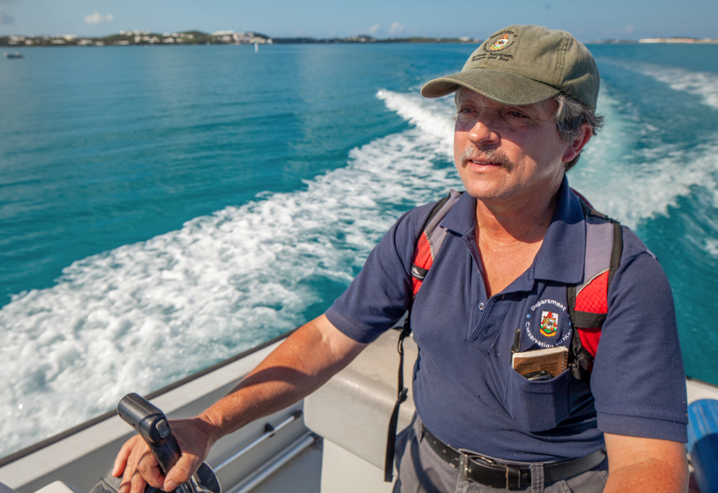 Ripples of white water cut through Bermuda's turquoise blue water, following a motorboat captained by Jeremy Madeiros. He wears a blue polo shirt, baseball cap, and gray moustache.