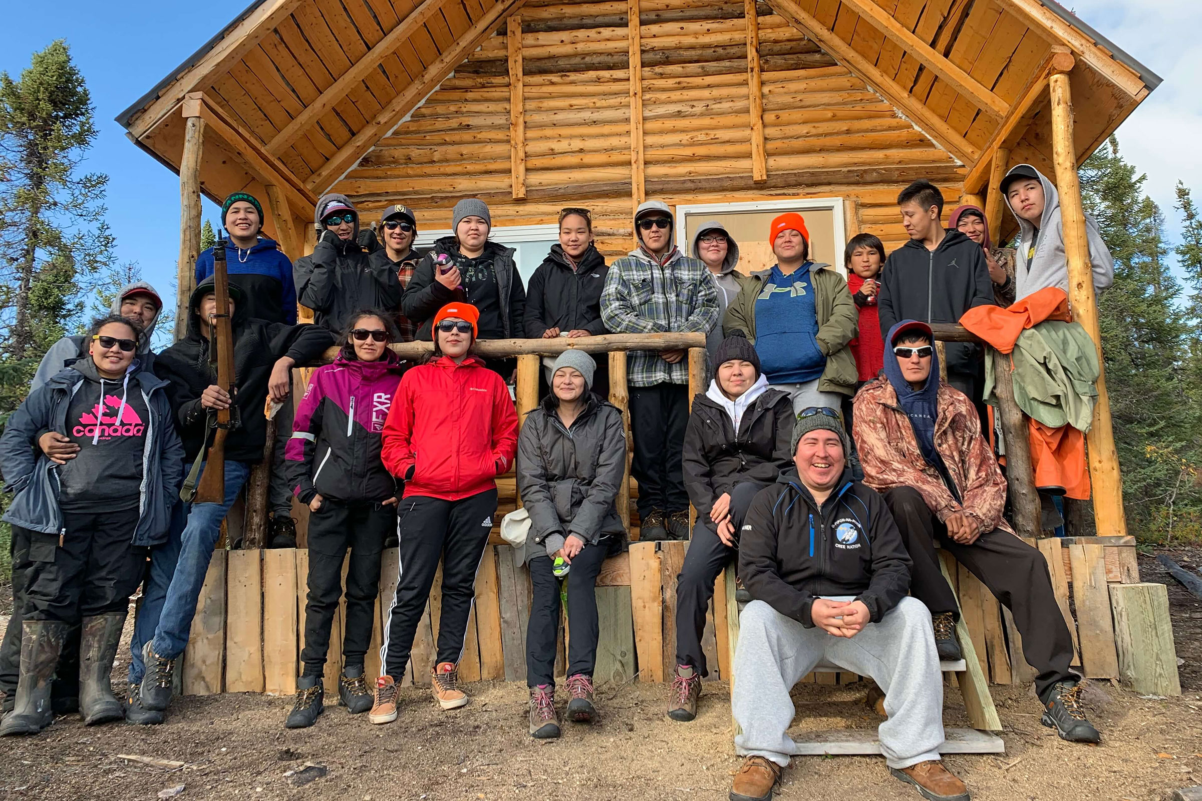 Fifteen Sayisi Dene First Nation, Northlands First Nation, O-Pipon-Na-Piwin Cree Nation, and Arviat, Nunavut youth, pictured standing and sitting outside of a log building on a sunny day, participated in the 2019 Tadoule Lake Youth Stewardship Summit, which focused on the cultural and environmental importance of the Seal River Watershed.